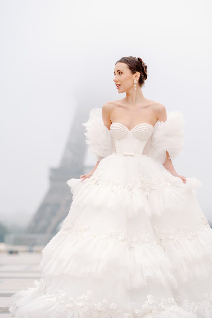 Paris pre-wedding session in front of the Eiffel Tower while it is covered in fog with bride in full train layered gown with separate off the shoulder full puffed sleeves and groom in a navy blue suit photographed by Italy destination photographer